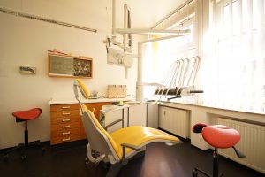 Einblick in ein Behandlungszimmer mit gelben Behandlungsstuhl und großer Fensterfront