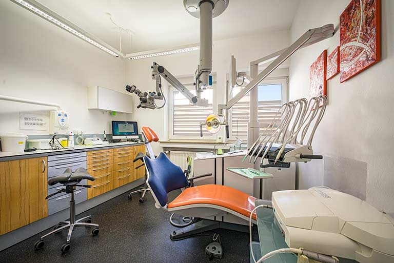 Kompleter Einblick in ein Behandlungszimmer mit orange-blauem Behandlungsstuhl, Tresen mit Computer im Eck und ein drei-teiliges rotes , abstraktes Bild an der Wand