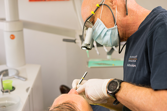 Zahnarzt W. Markert ist mit Mundschutz und Stirnlampe in Seitenansicht zu sehen, wie er den Mund eines Patienten inspiziert
